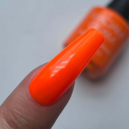 Close-up of a hand with an orange painted fingernail and a blurred nail polish bottle in the background.