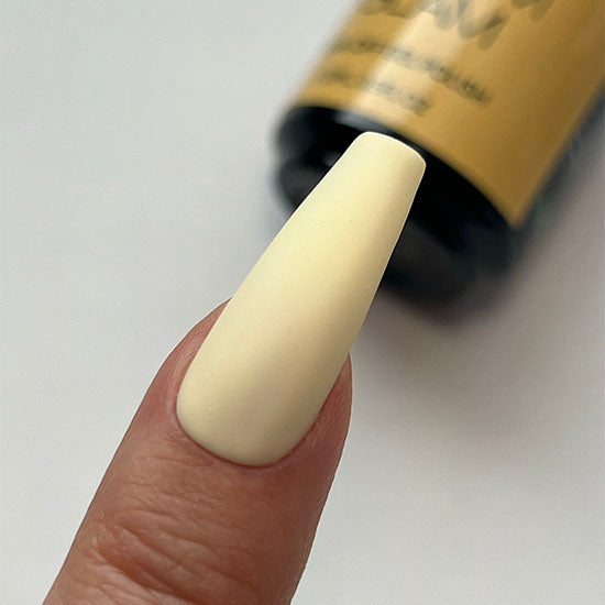 Nail with beige polish held by a finger, with a blurred bottle of nail polish in the background.