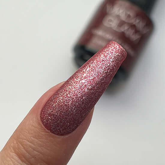 Close-up of a glittery pink nail polish on a finger with a blurred bottle in the background.