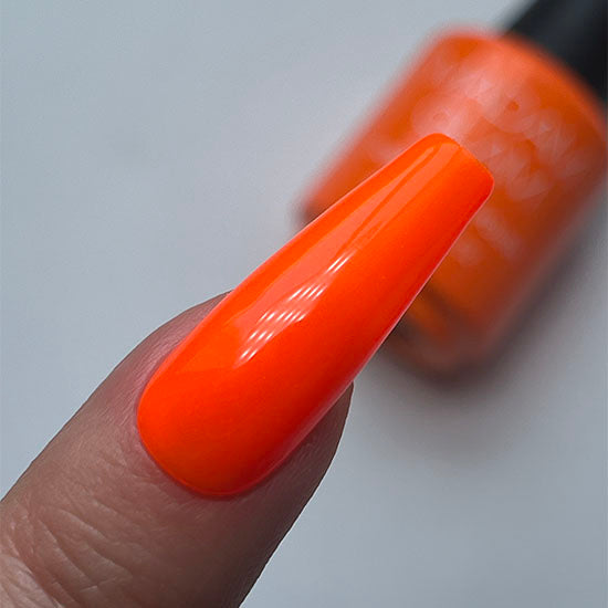 Close-up of a hand with an orange painted fingernail and a blurred nail polish bottle in the background.