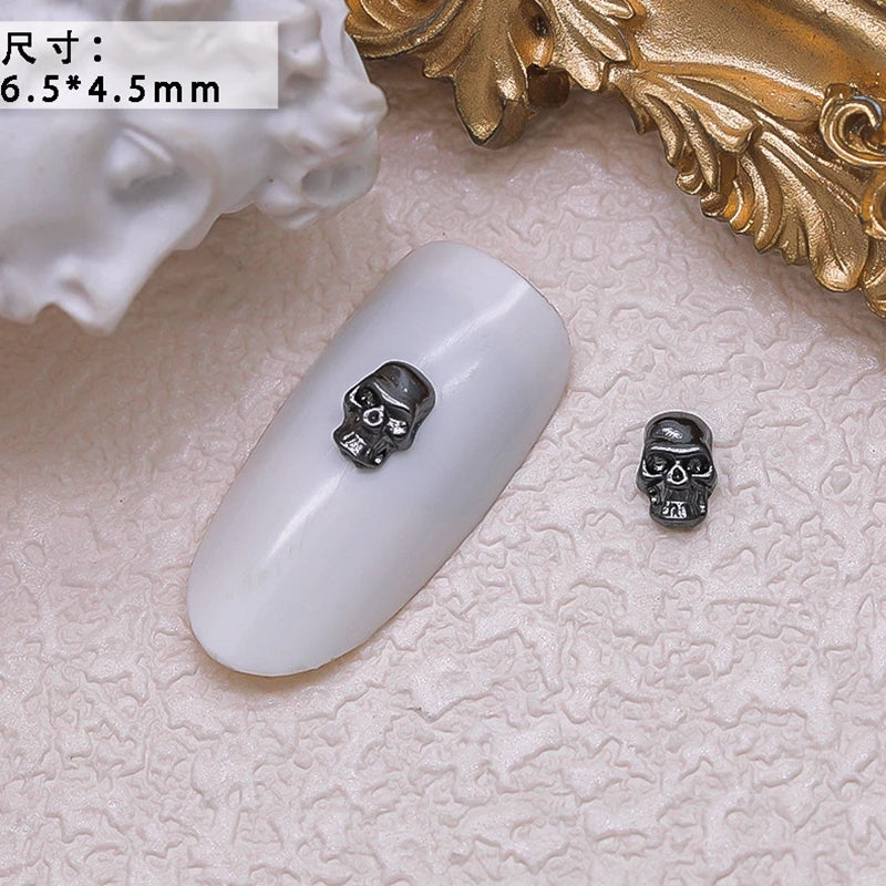 Small skull-shaped nail art decorations on a white nail with a gold decorative object in the background.