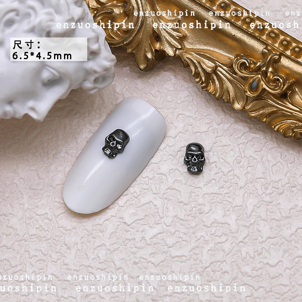 Small skull-shaped nail art decorations on a white surface with gold decorative frame.